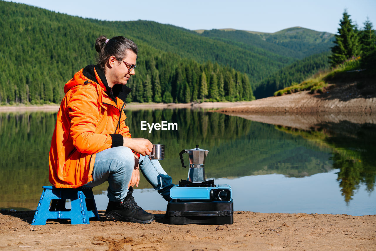 Side view of a man sitting by camping stove by the lake against mountains