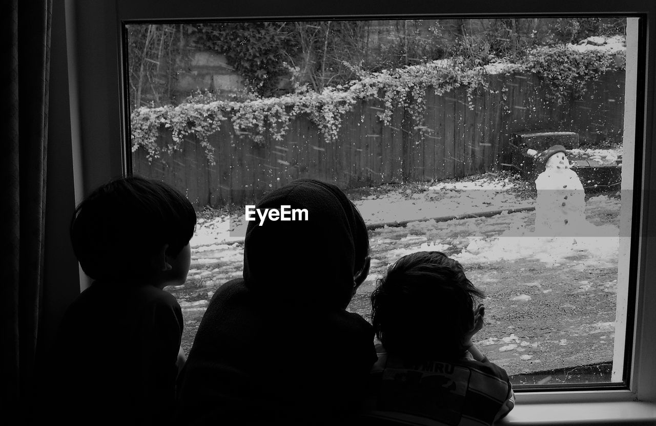 Rear view of siblings looking at snowman through window at home