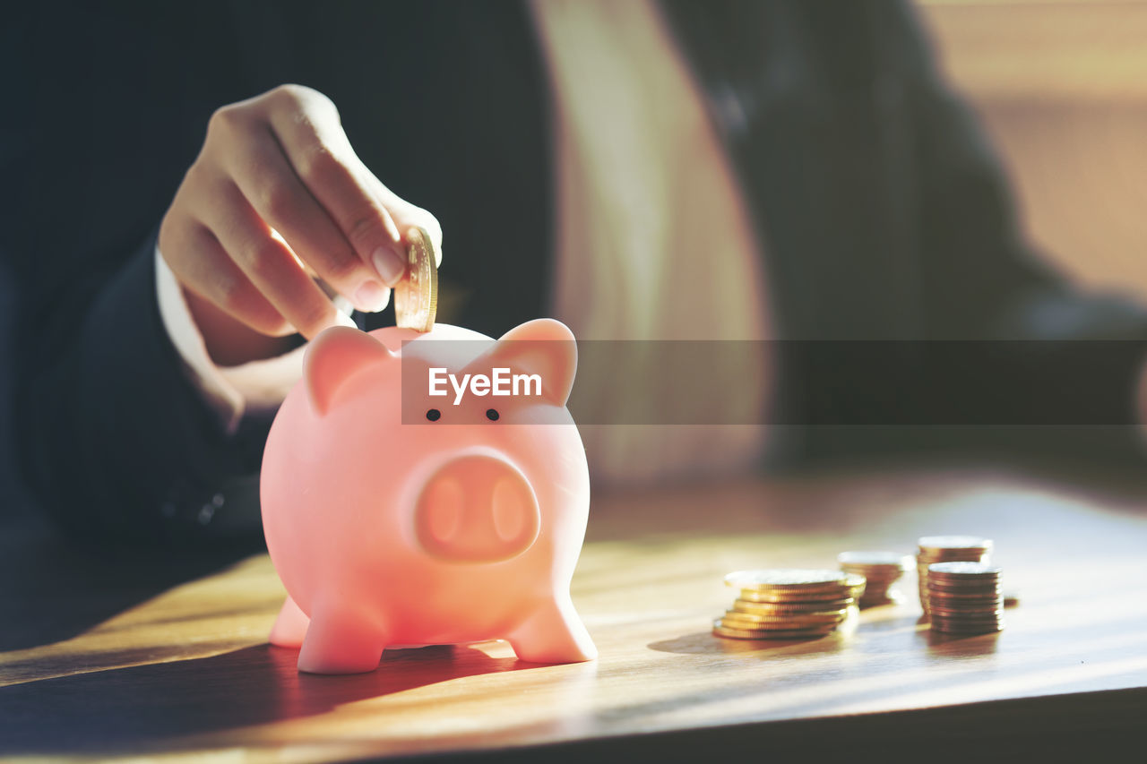 Midsection of businesswoman inserting coins in piggy bank on table