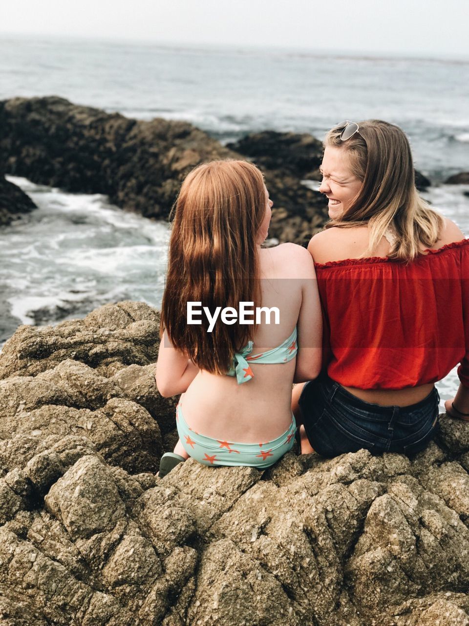 REAR VIEW OF WOMEN SITTING ON ROCK BY SEA