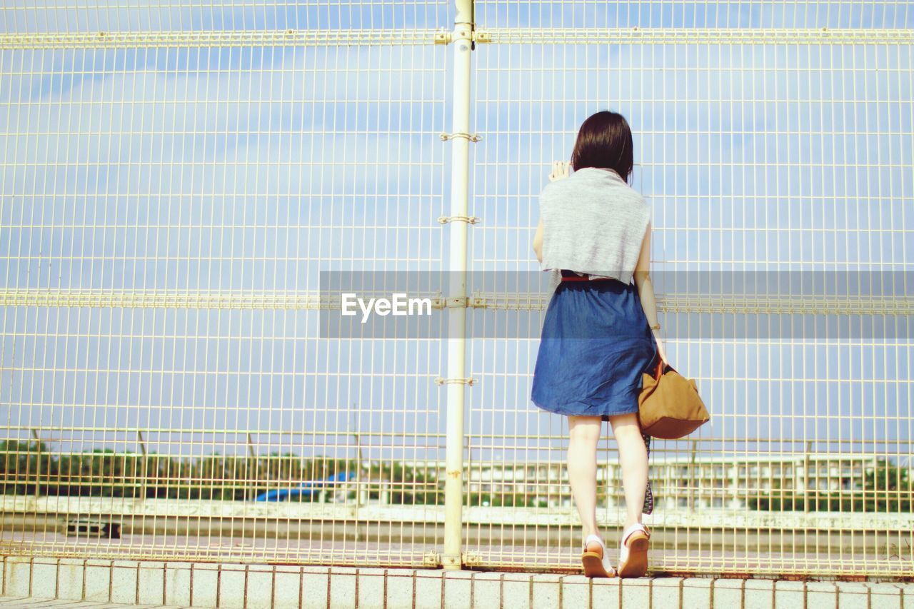 Rear view of girl looking through metal fence against sky