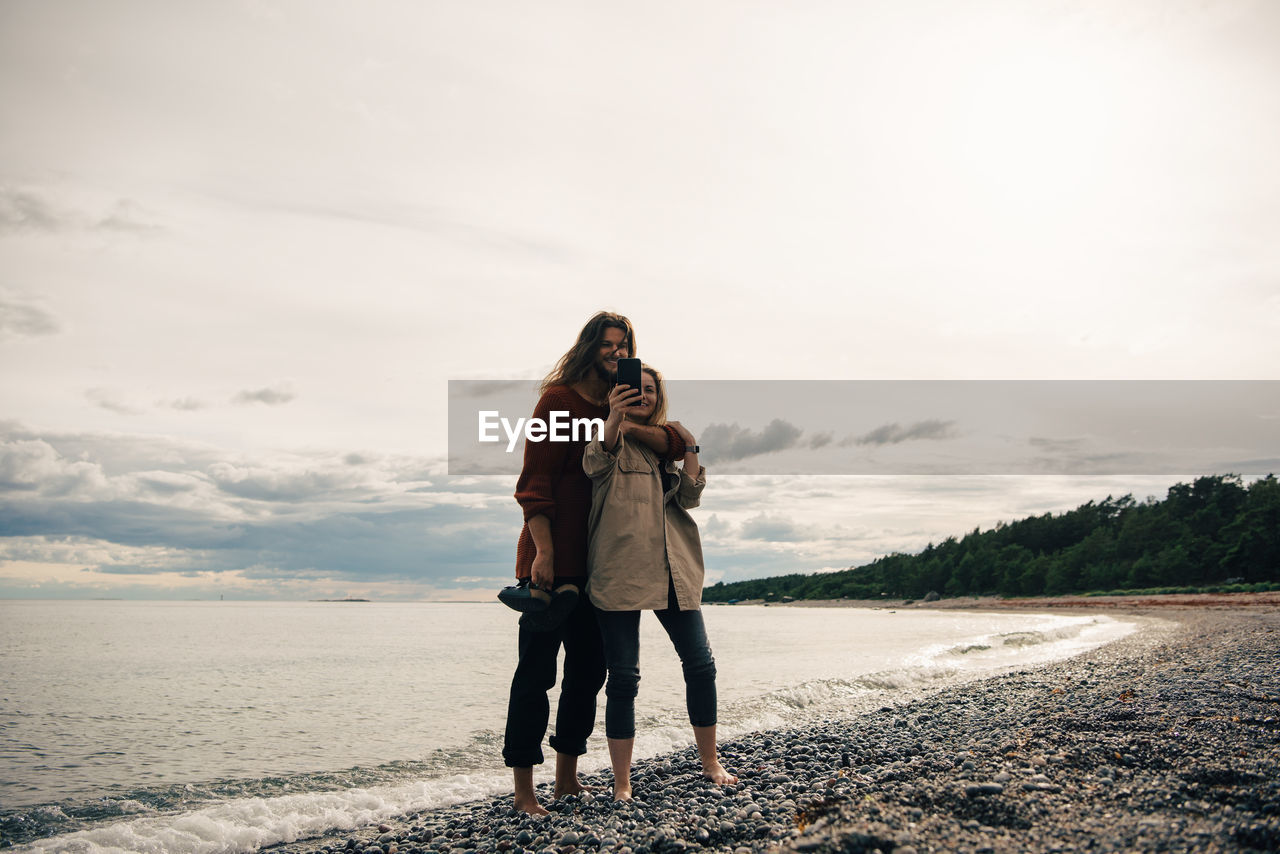 Full length of couple taking selfie on smart phone at beach against sky