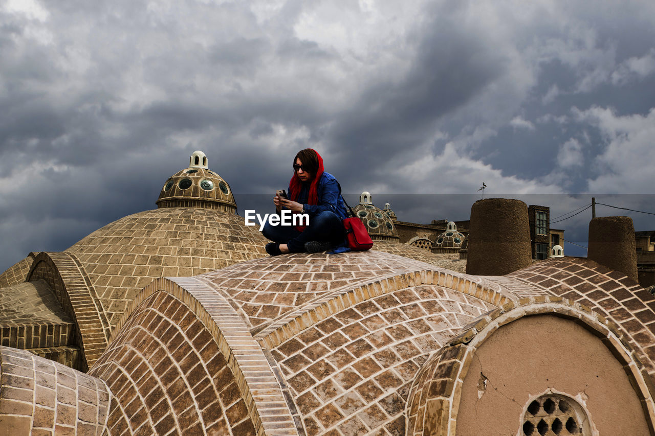 Woman sitting on built structure against cloudy sky