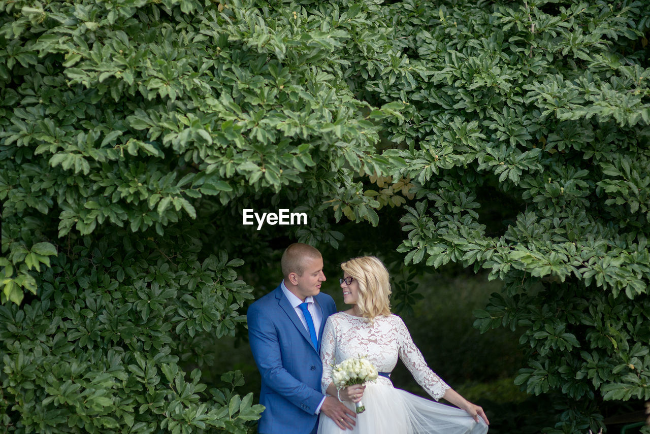 Bridal couple looking at each other while standing amidst tree branches at park