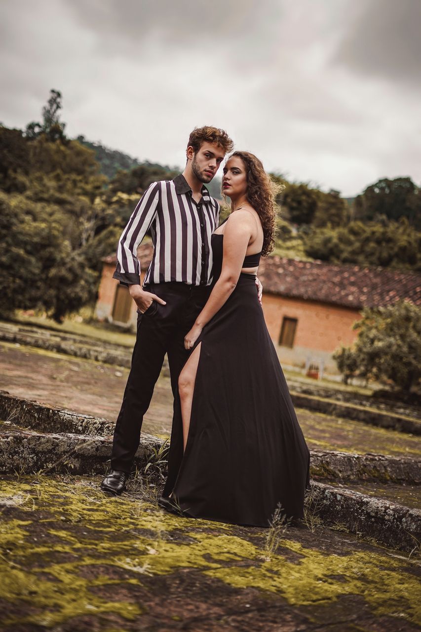 PORTRAIT OF YOUNG COUPLE STANDING OUTDOORS AGAINST SKY