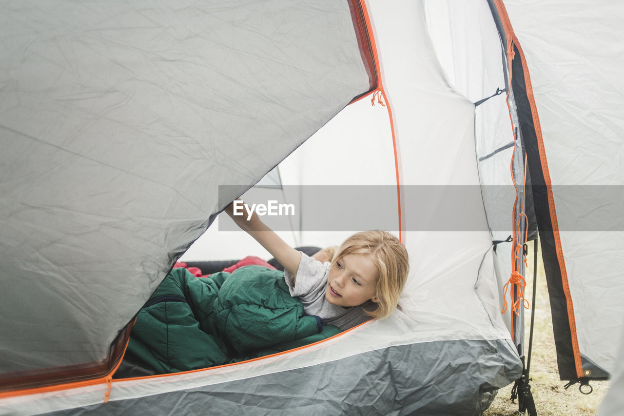 Girl in sleeping bag peeking through tent at camping site