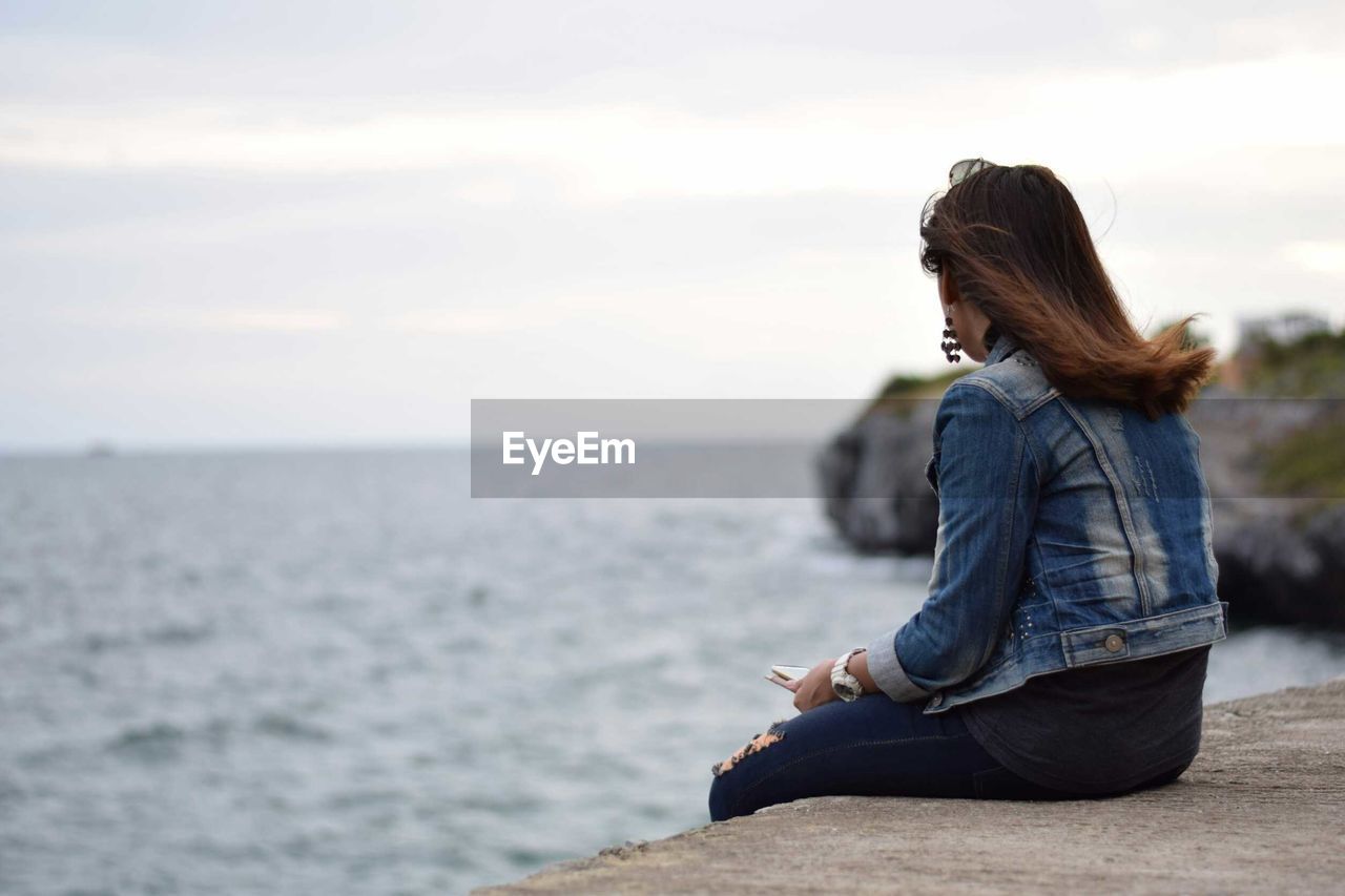 Side view of woman looking at sea while sitting on retaining wall against sky