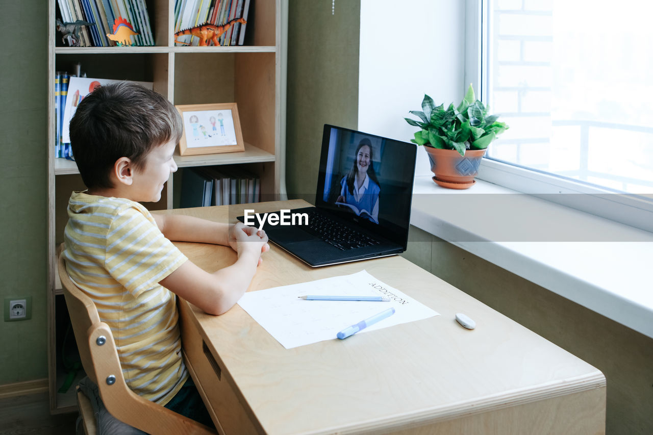 7 years old boy sitting by desk with laptop doing writing task during online lesson. back view