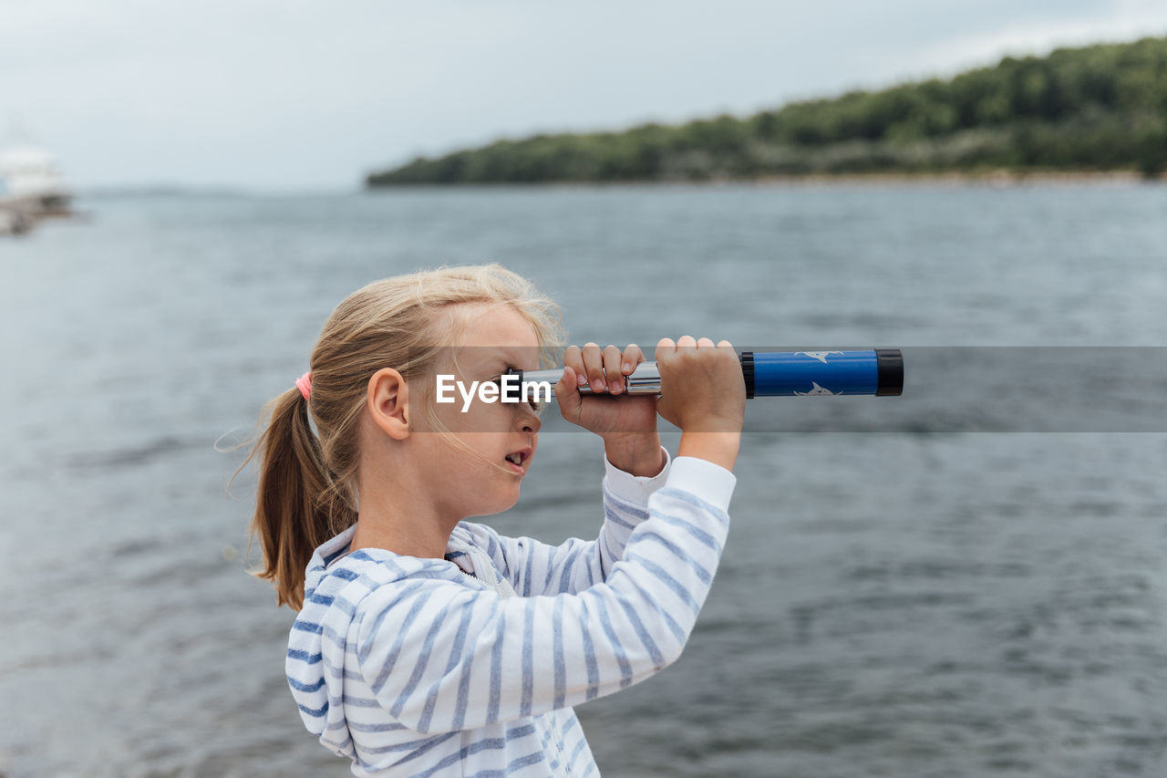 Side view of girl looking through telescope while standing by sea against sky