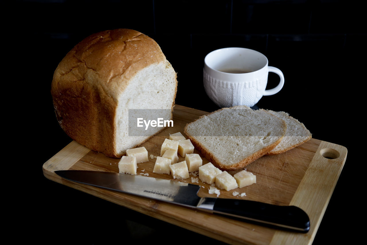 Bread and knife on cutting board by coffee cup over black background