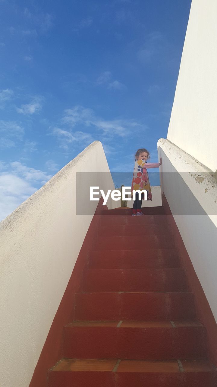 LOW ANGLE VIEW OF GIRL STANDING ON STAIRCASE AGAINST SKY