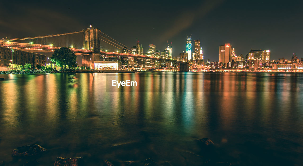 ILLUMINATED BRIDGE OVER RIVER BY BUILDINGS AGAINST SKY