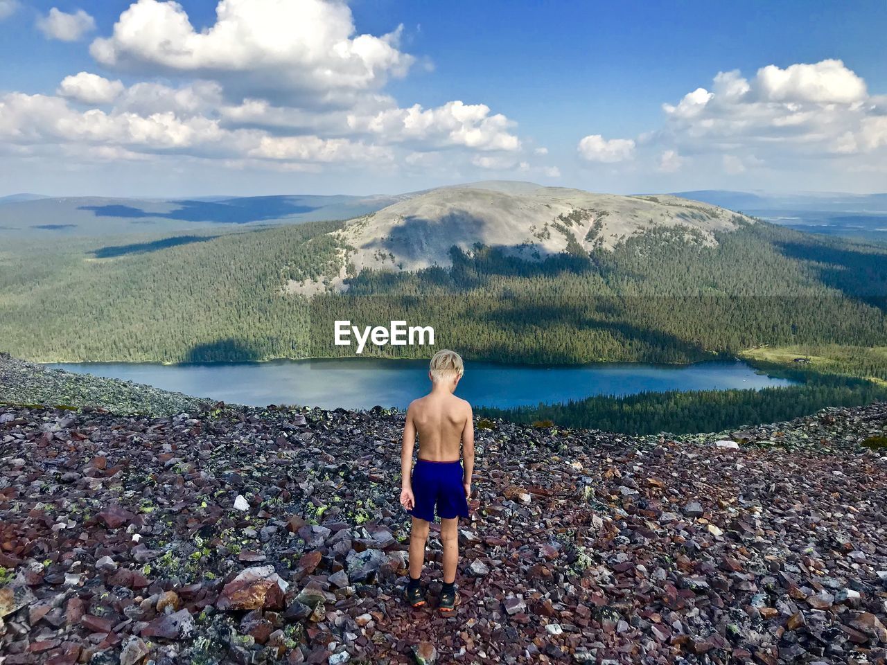 Rear view of shirtless boy looking at lake and mountains against sky