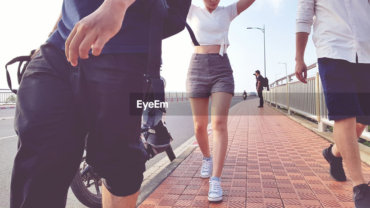 Low section of men and woman walking on footpath against sky