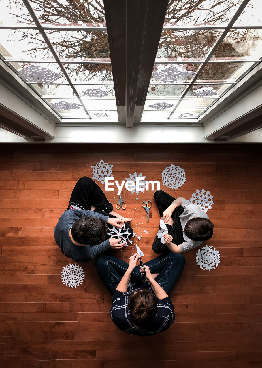 Woman and two boys sitting on floor cutting paper snowflakes. .