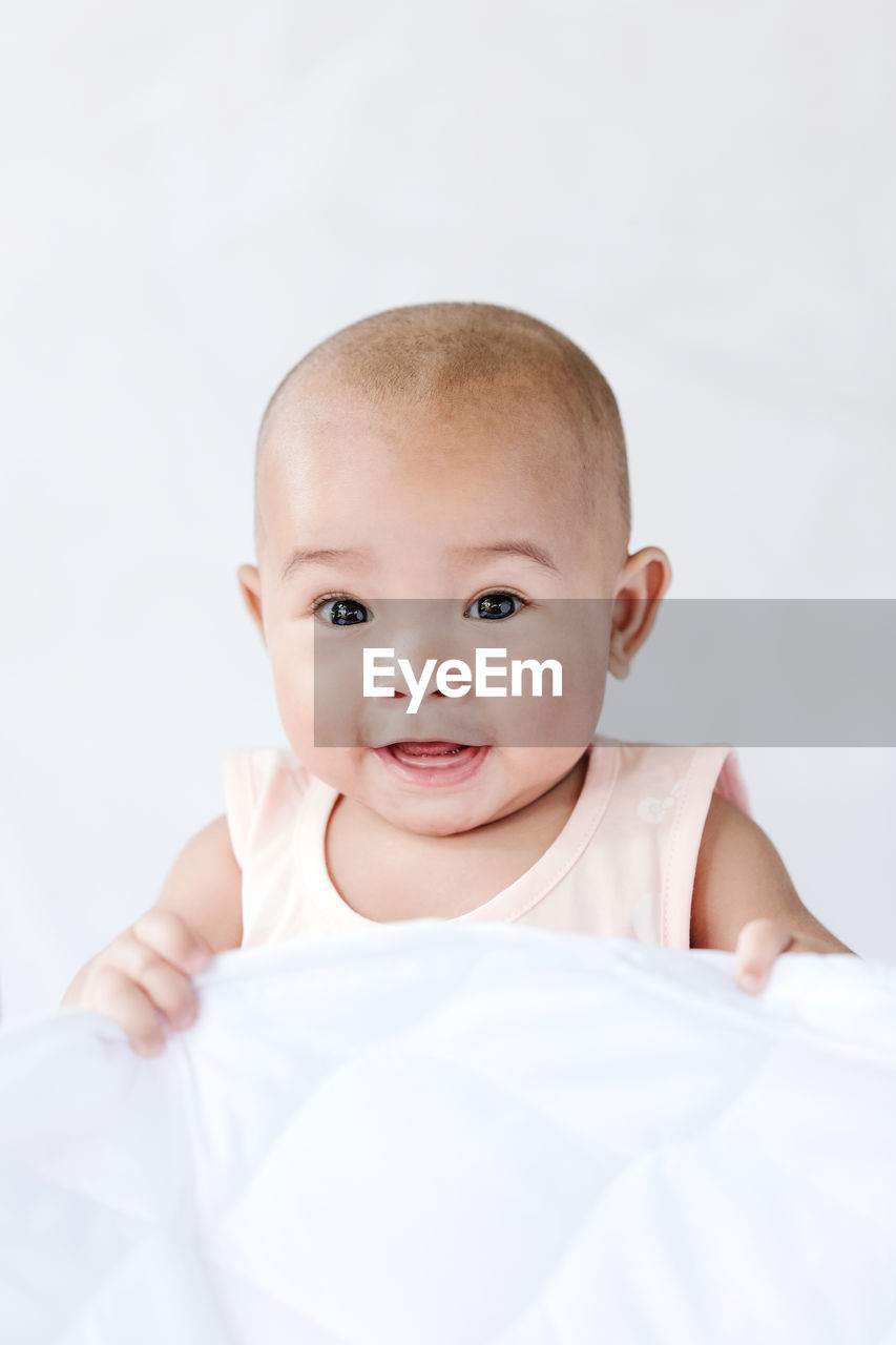 Close-up portrait of cute baby girl sitting on bed against wall at home