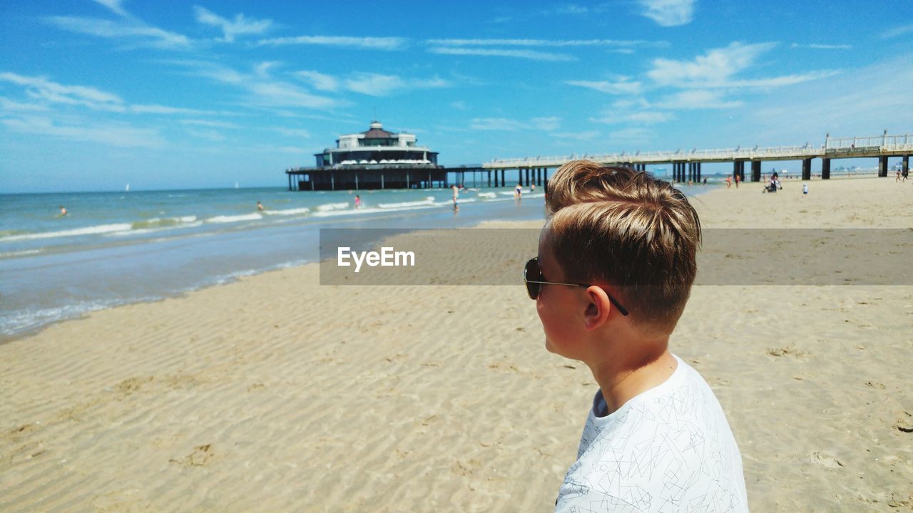 Side view of boy standing at beach against blue sky