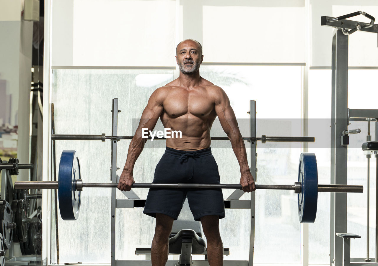 Portrait of shirtless man holding barbell standing in gym