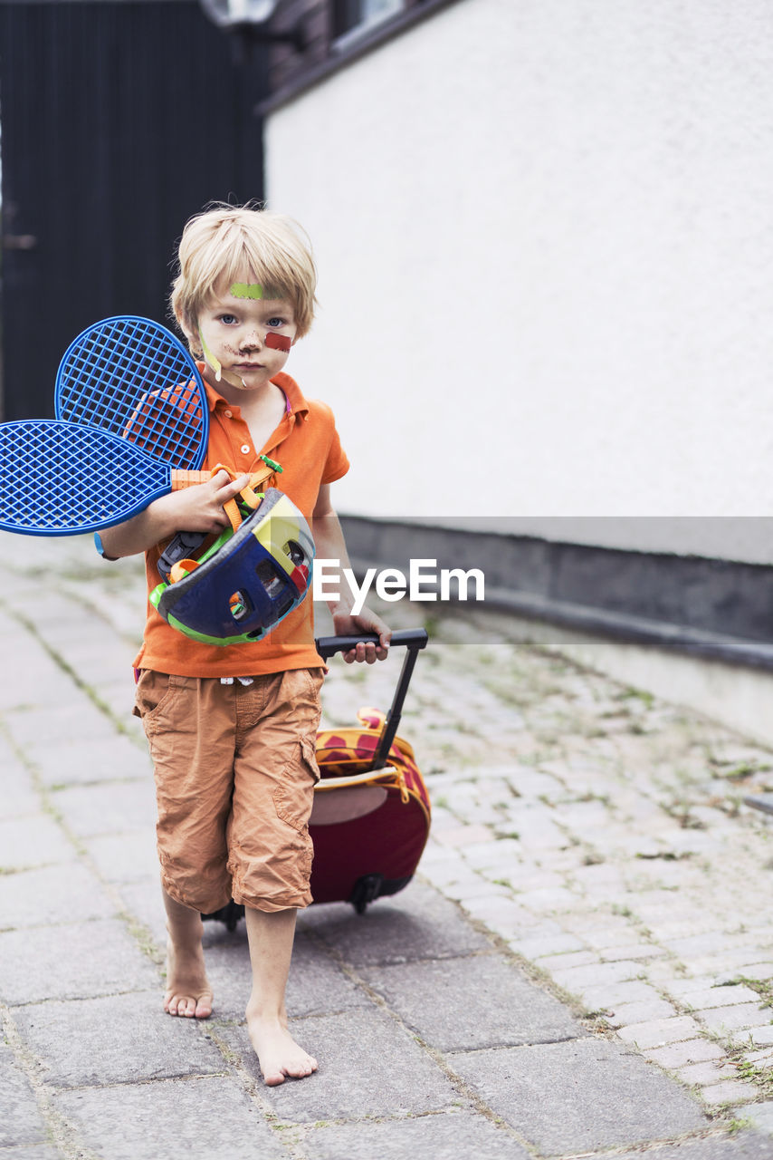 Full length portrait of boy pulling luggage while holding tennis rackets and helmet on street