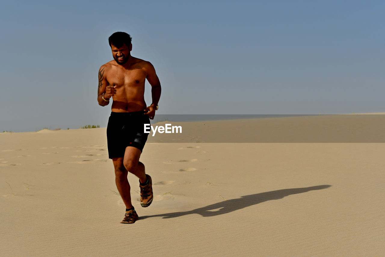Full length of shirtless young man running on sand at desert during sunny day