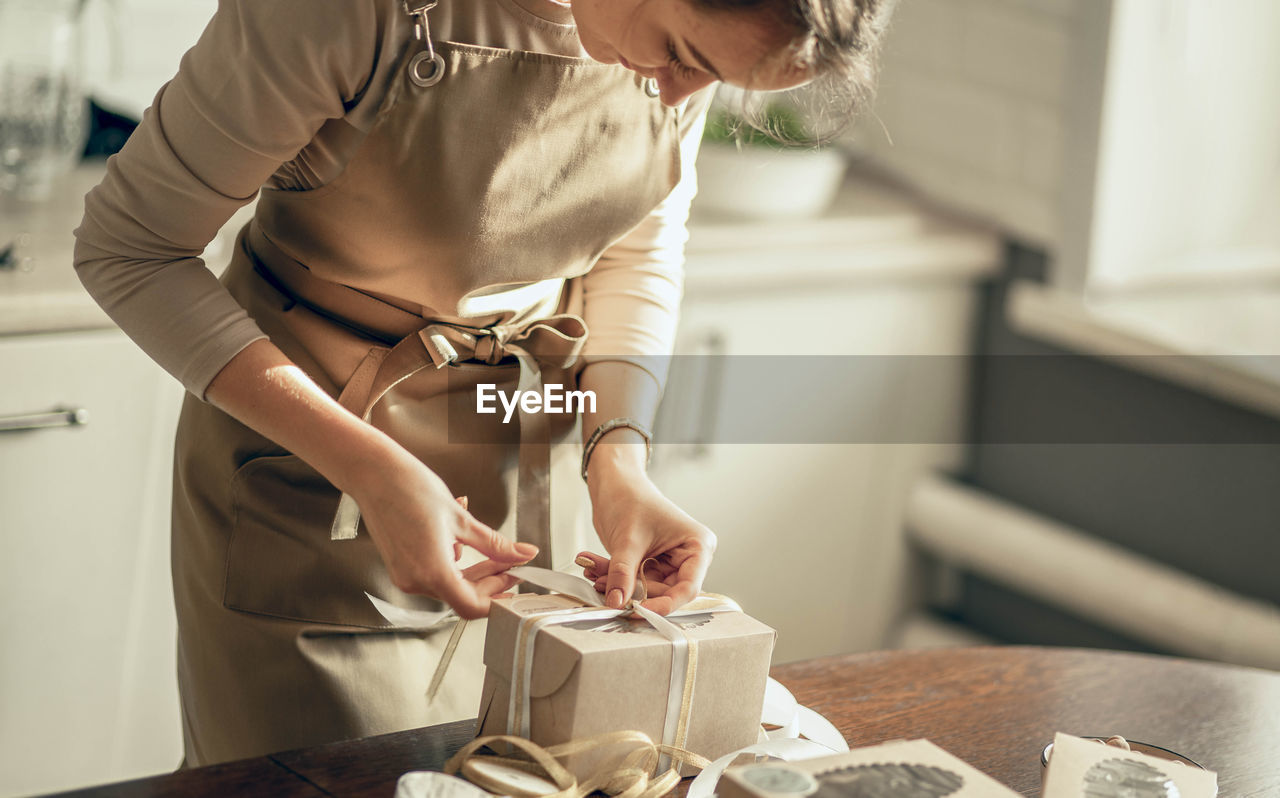 Woman bakery shop owner ties ribbon on box for customer order. chef baking pastry, cake in kitchen