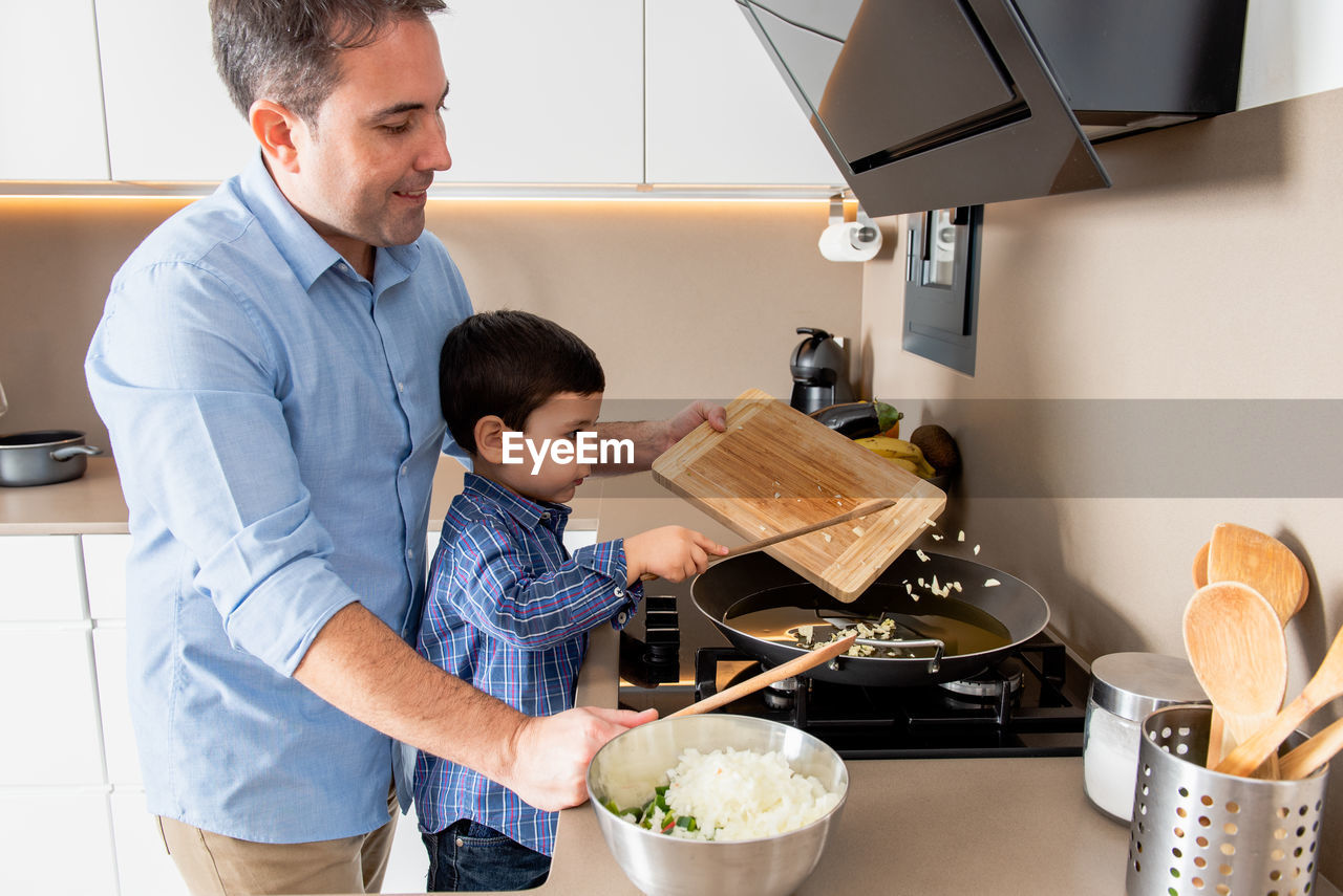 Side view cute concentrated boy stirring garlic on frying pan while learning to cook with father