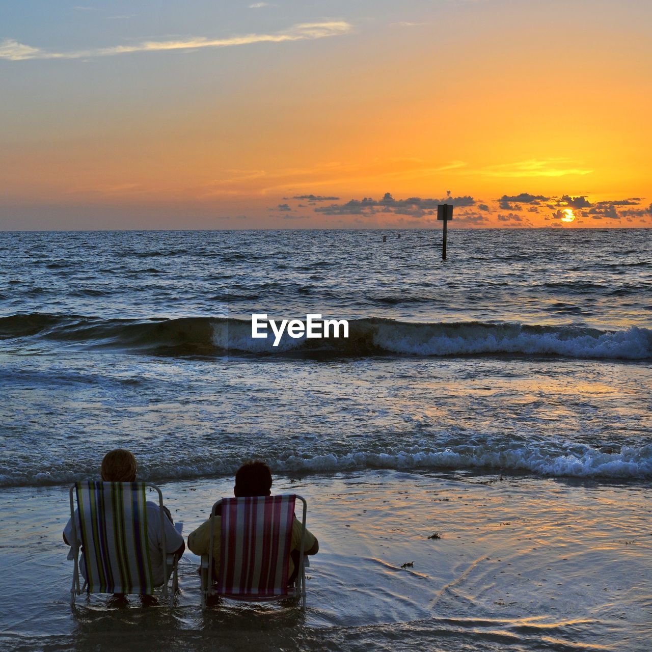 Rear view of friends sitting on deck chairs at beach against sky during sunset