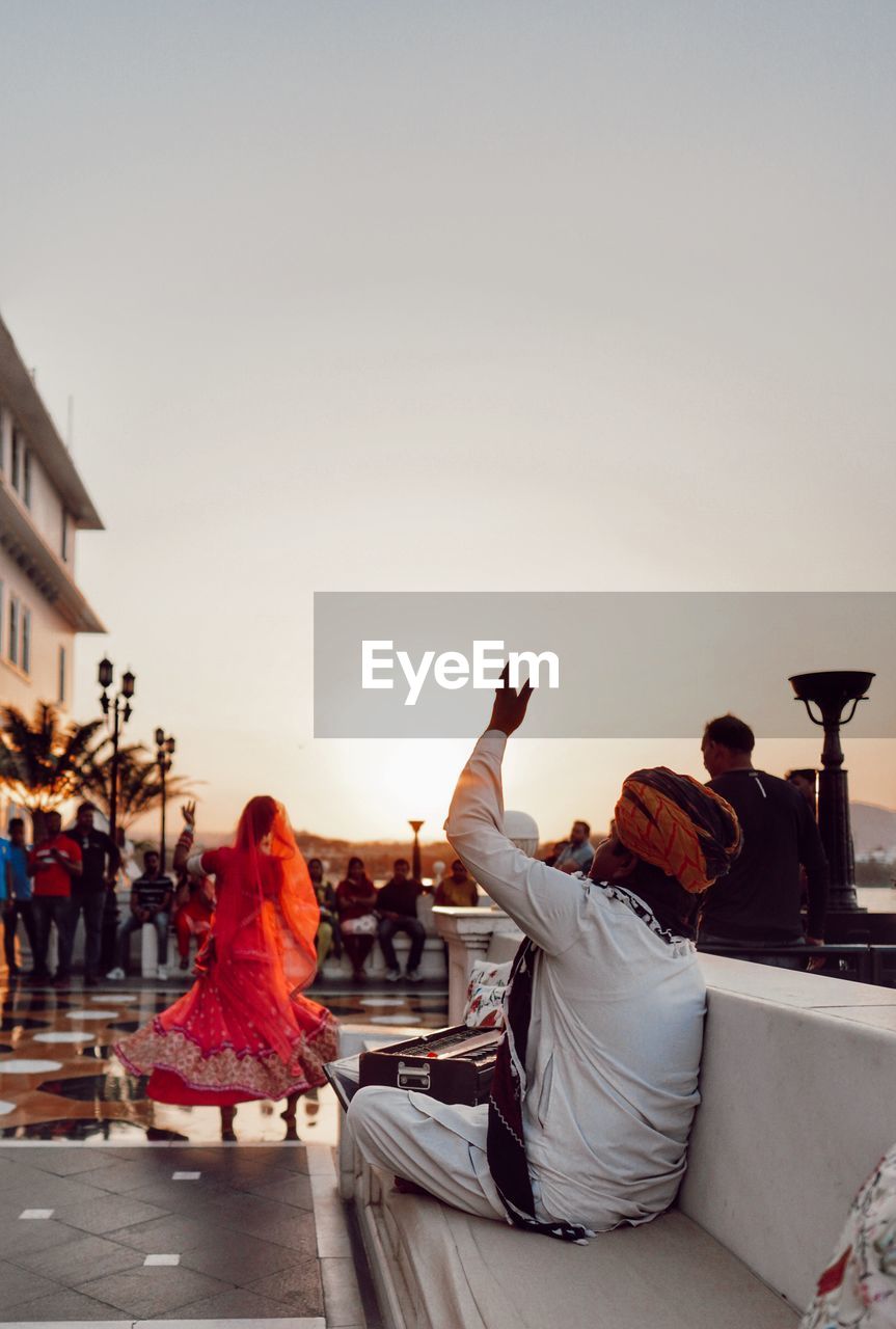 REAR VIEW OF PEOPLE SITTING AGAINST SKY DURING SUNSET