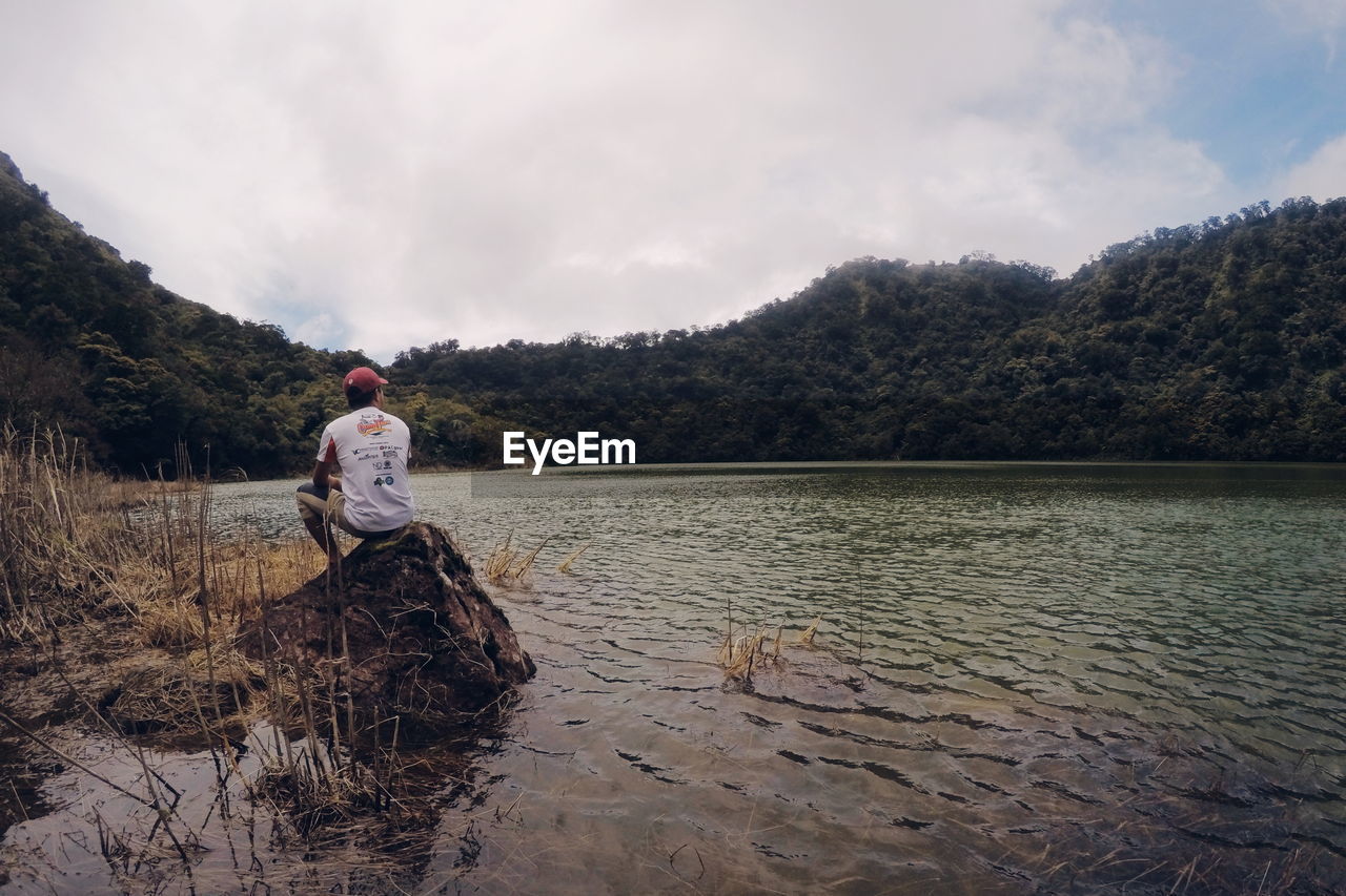 Rear view of man looking at lake while sitting on rock against sky