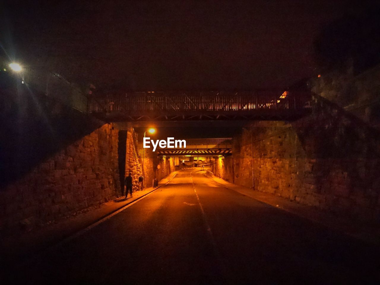 ILLUMINATED ROAD AMIDST BRIDGE AT NIGHT