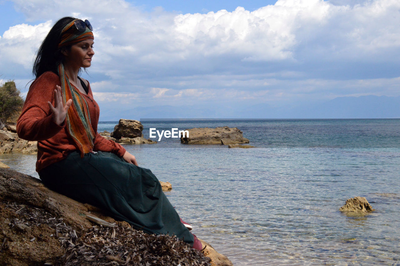 Side view of young woman meditating while sitting on rock at beach