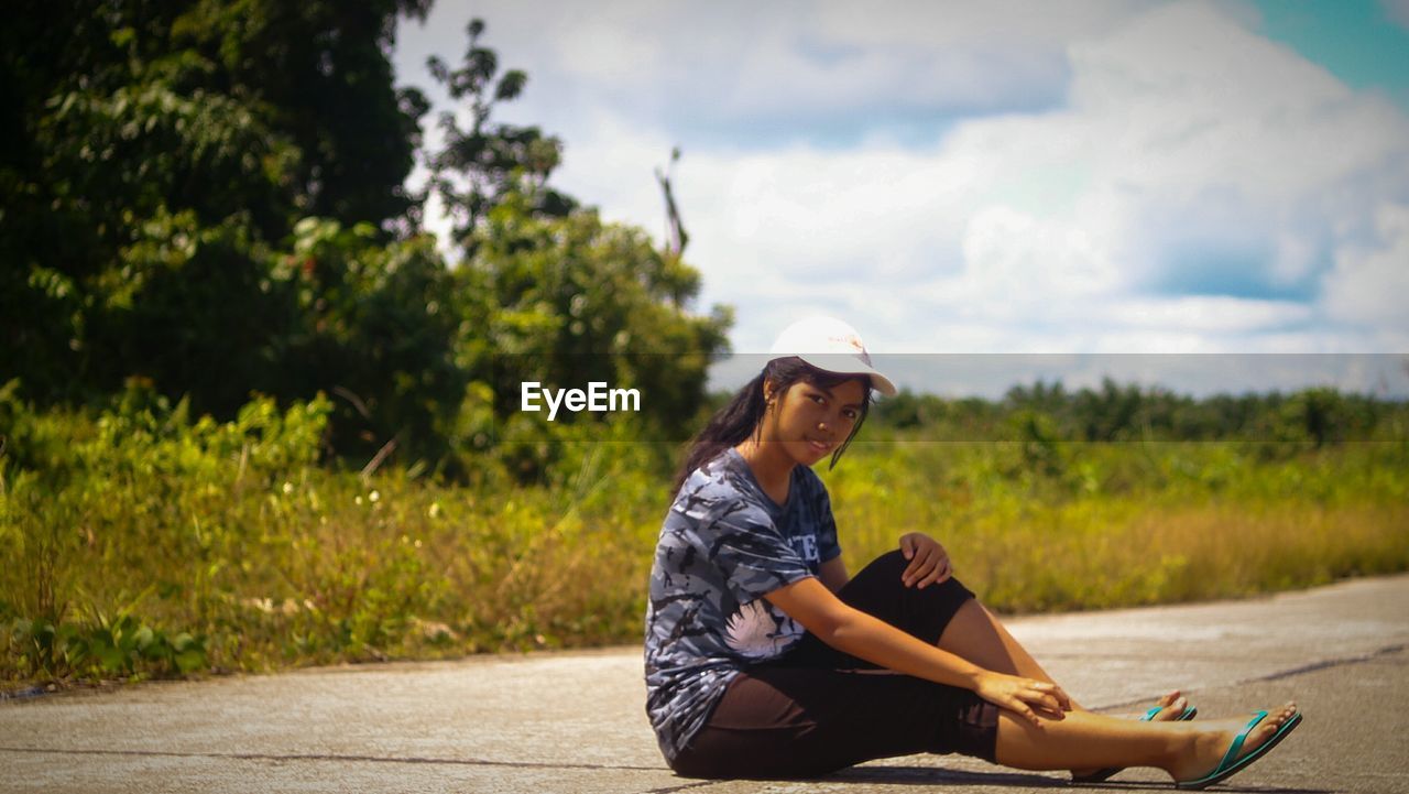 Portrait of young woman sitting on road by trees against sky