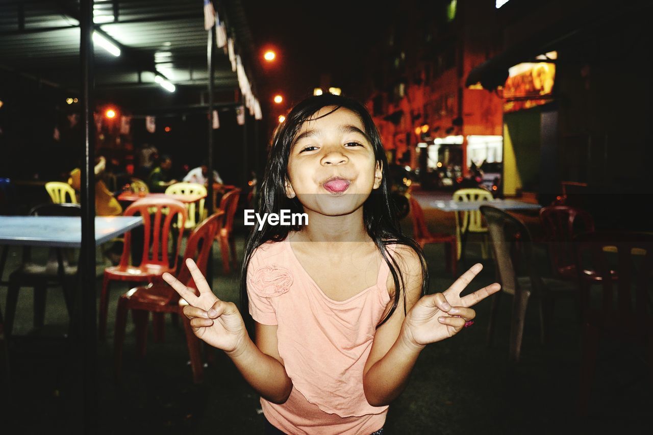 Portrait of girl sticking out tongue while gesturing peace sign in city at night