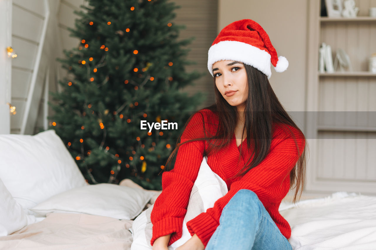 Young sad asian woman in red sweater and santa hat on bed in room with christmas tree at home