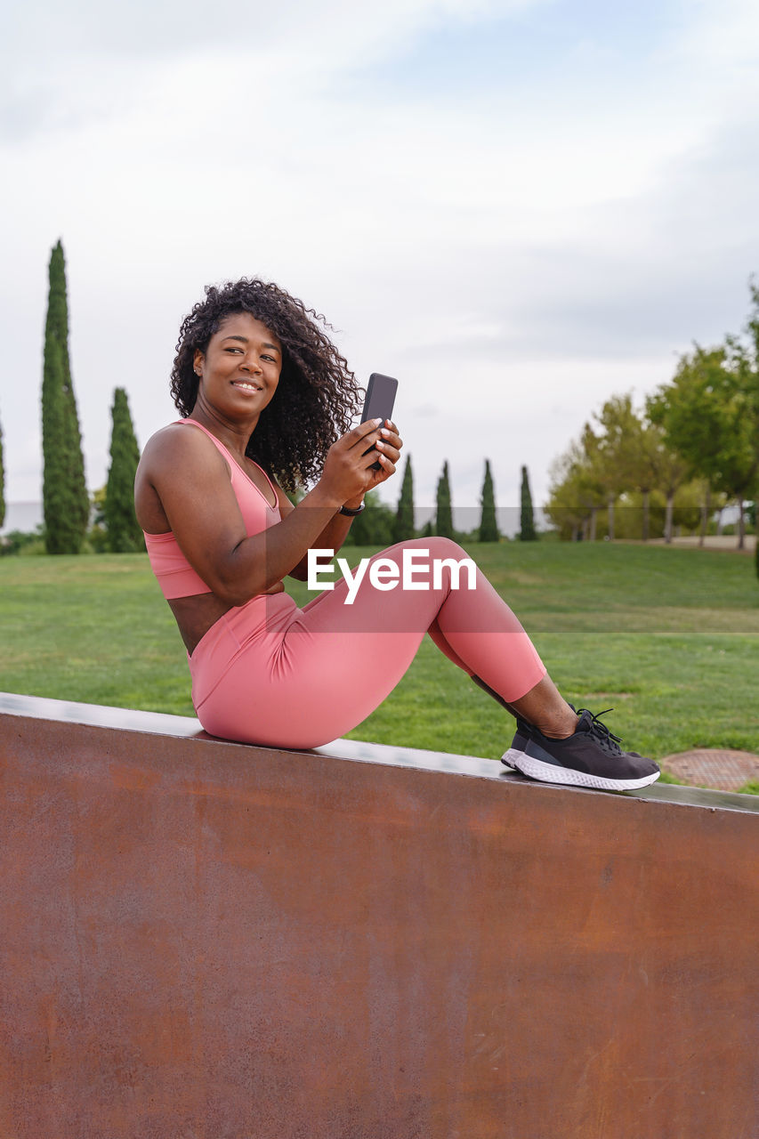 PORTRAIT OF YOUNG WOMAN USING PHONE WHILE SITTING ON CAMERA