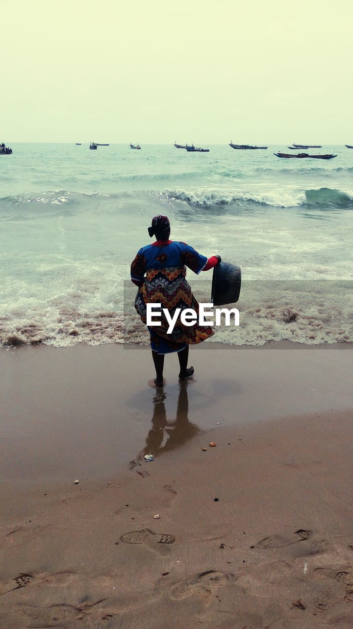 Rear view of woman holding basket while standing on shore against clear sky