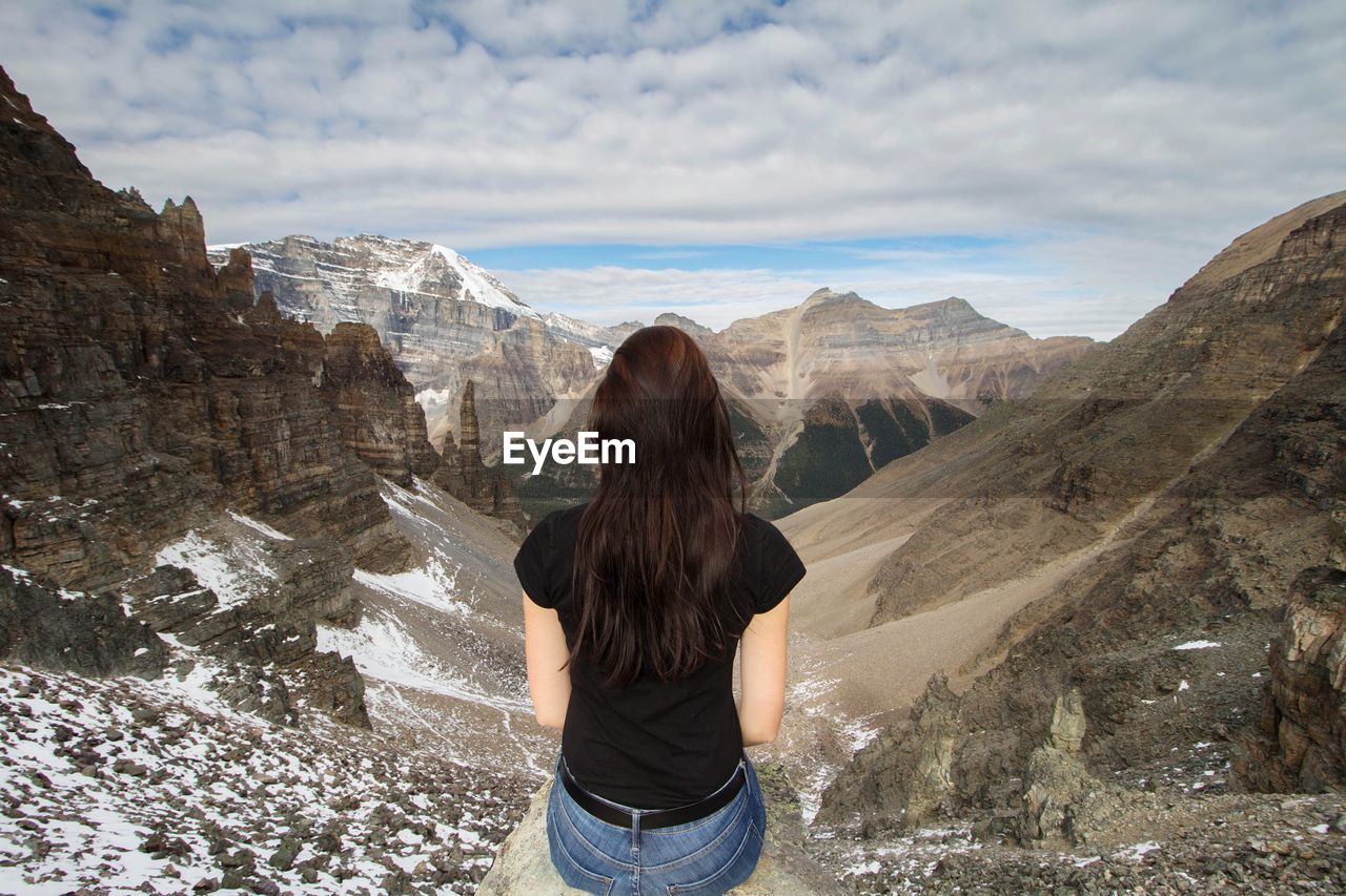 Rear view of woman sitting on cliff against mountains during winter