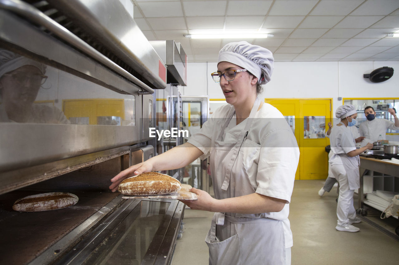 Side view of focused female baker taking out freshly baked bread from hot oven while learning baking in baker school