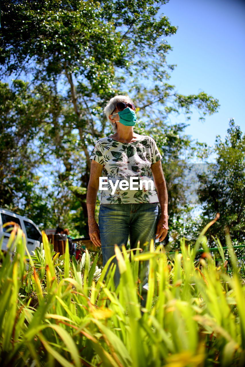 Low angle view of elderly woman standing by tree on field wearing mask