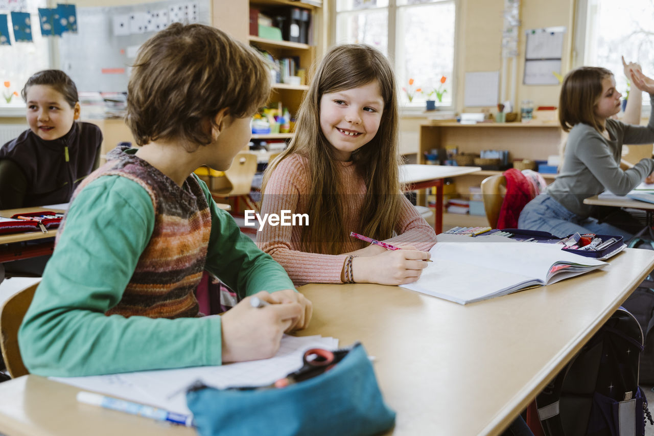 Smiling girl talking to male friend while sitting at desk in classroom