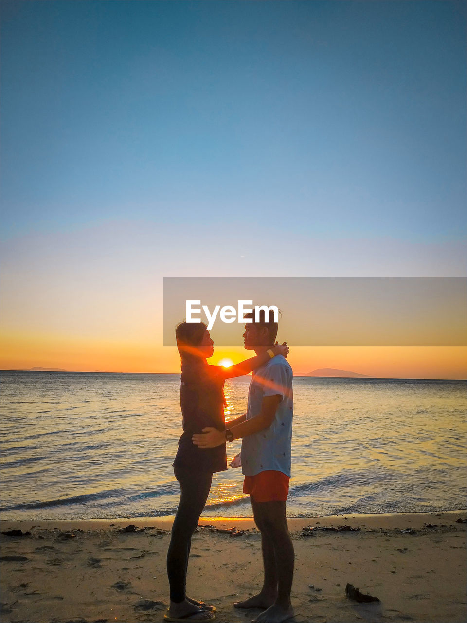 MEN STANDING ON BEACH AGAINST SEA DURING SUNSET