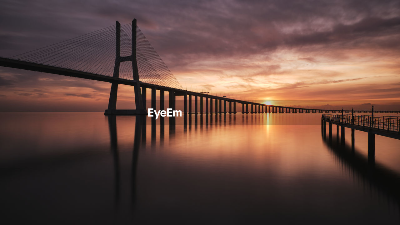 Silhouette bridge over sea against sky during sunset