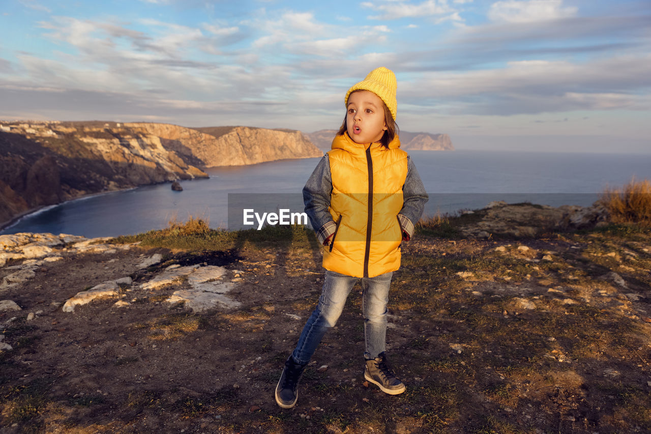 Boy child in a yellow hat and vest stands on top of a mountain by the sea in autumn