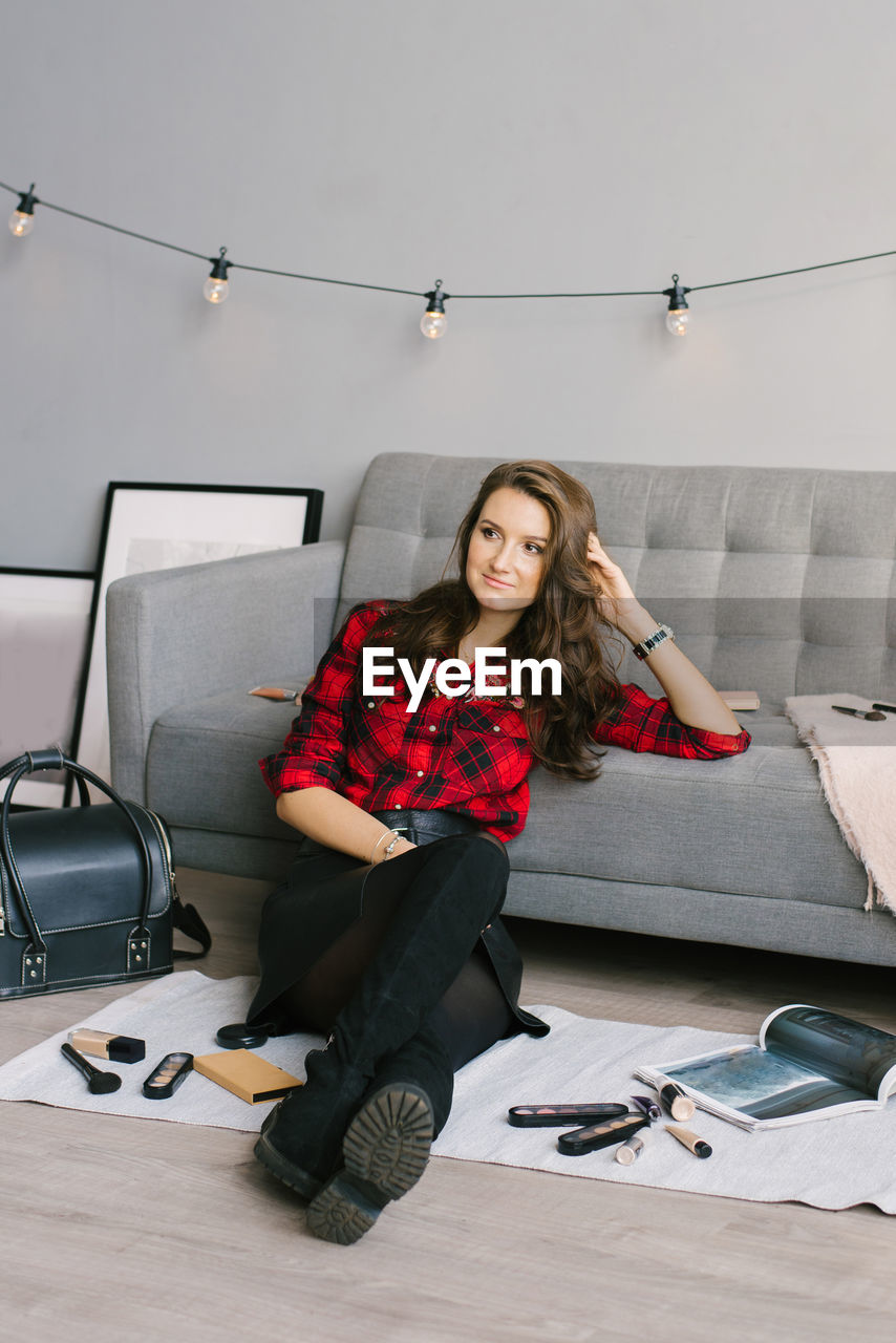 Young female makeup artist is sitting on the sofa floor next to randomly scattered brushes, makeup