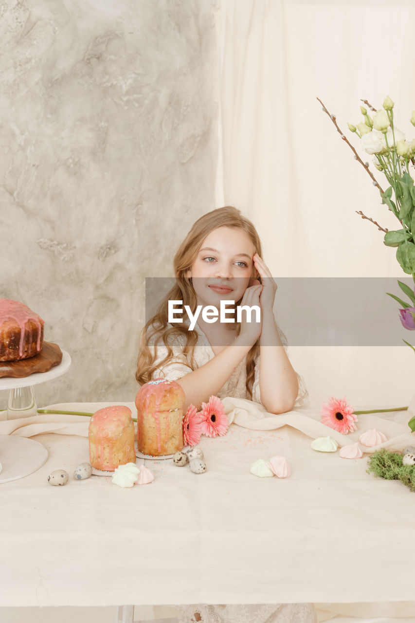 A girl with long hair in a light dress is sitting at the easter table with cakes, spring flowers 