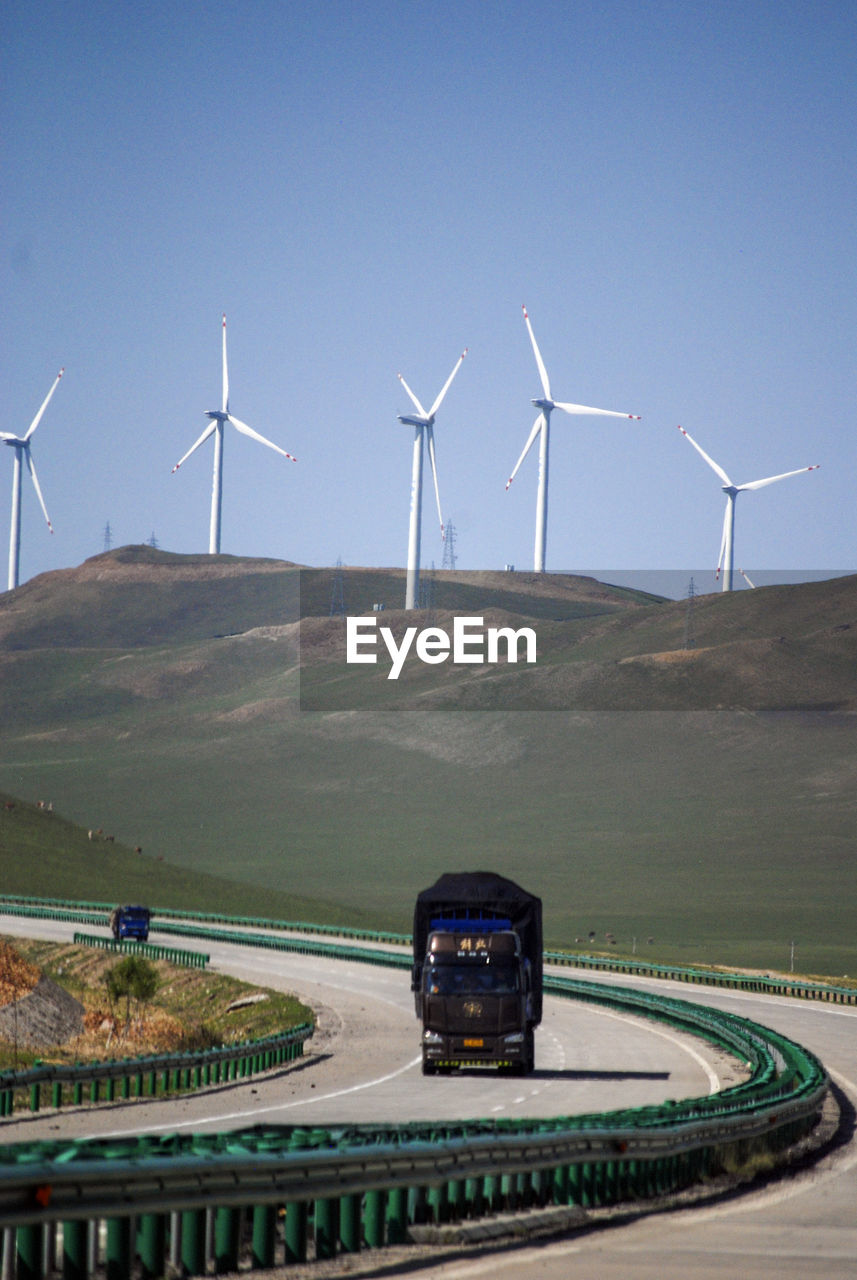 There is a highway at the foot of the mountain. there is a white electric windmill on the mountain