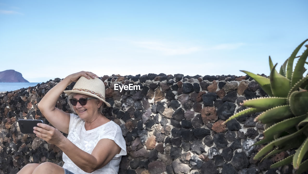 Smiling senior woman taking selfie while sitting by retaining wall