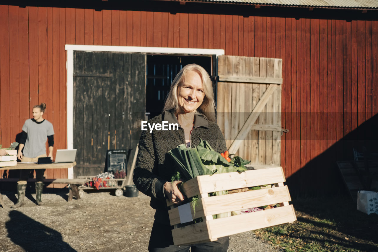 Portrait of smiling mature woman carrying crate full of vegetables with barn in background