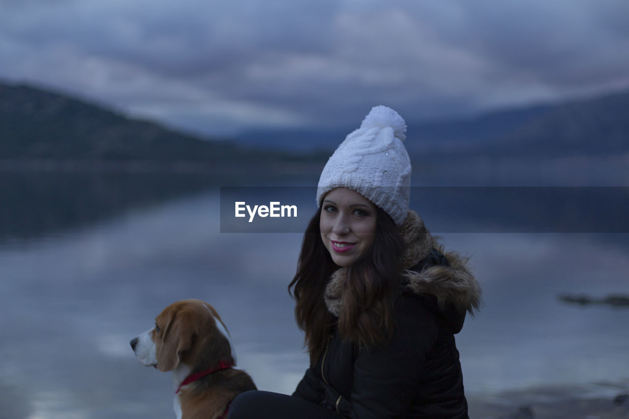 Portrait of smiling woman with beagle sitting by lake against sky during winter