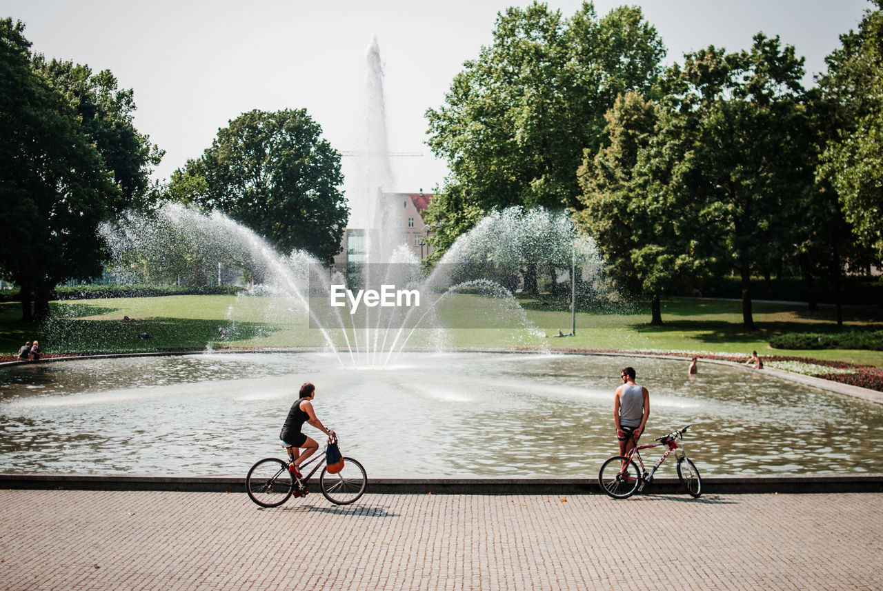 Woman cycling while man leaning on bicycle by fountain at park